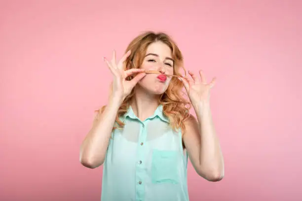 funny amusing playful girl fooling around with her hair. portrait of a young carefree woman making a mustache with her locks on pink background.