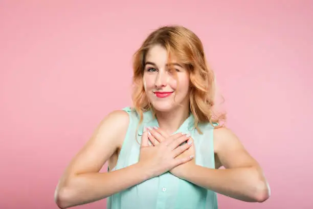 flattered and happy pretty blond girl. smiling and pressing both hands to her heart. sweet reaction and pleasant feelings concept, young beautiful woman on pink background.