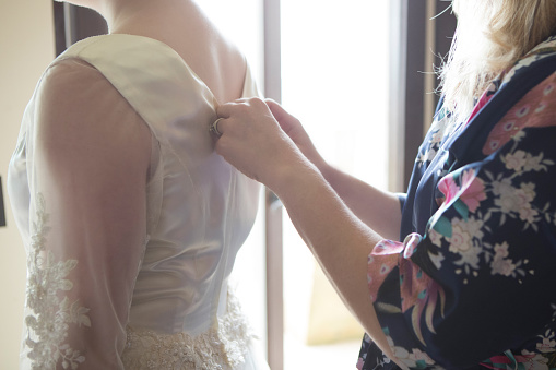 In this closeup, both are unrecognizable as a mother of the bride zips her daughter's wedding dress in the back.  There is sunlight streaming through a window in the background.