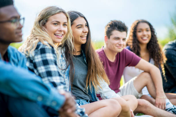 Young adult friends laughing together on the grass A diverse group of young adults laugh as they sit next to each other on the green grass. junior level stock pictures, royalty-free photos & images