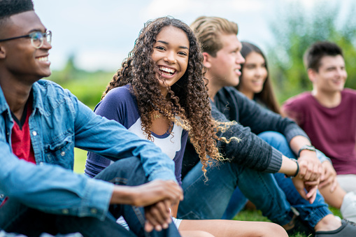 A diverse group of teens sits outside on the grass in the summertime. A girl is laughing playfully and looking at the camera, while her friends smile and look into the distance.