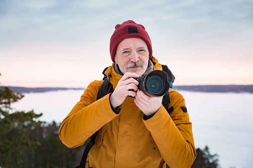 Portrait of mature man with grey beard exploring Finland in winter. Traveler Taking pictures on the top of rock. Beautiful view of northern landscape with frozen Baltic Sea and snowy islands.