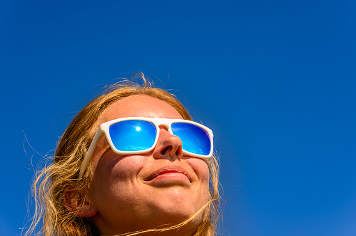 A blonde haired female model wearing sunglasses with blue lenses against a blue sky background.