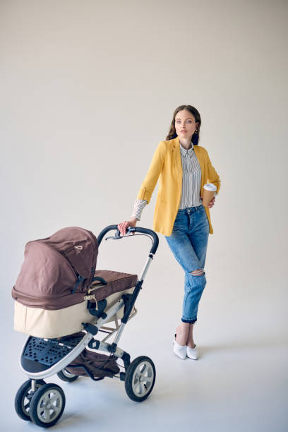 istock stylish young woman holding disposable coffee cup while standing with baby stroller and looking at camera on grey 1041667204