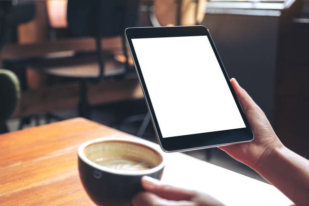 istock Mockup image of woman's hands holding black tablet pc with blank white screen while drinking coffee on wooden table in cafe 1041673948