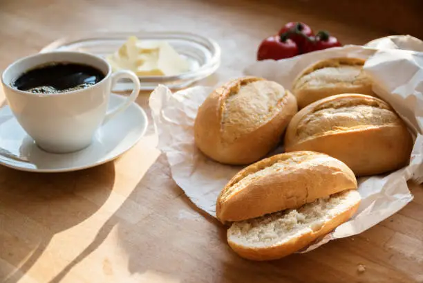 bread rolls or buns in a white paper bag, tomatoes, butter and a cup of coffee for breakfast on a wooden table, selected focus, narrow depth of field