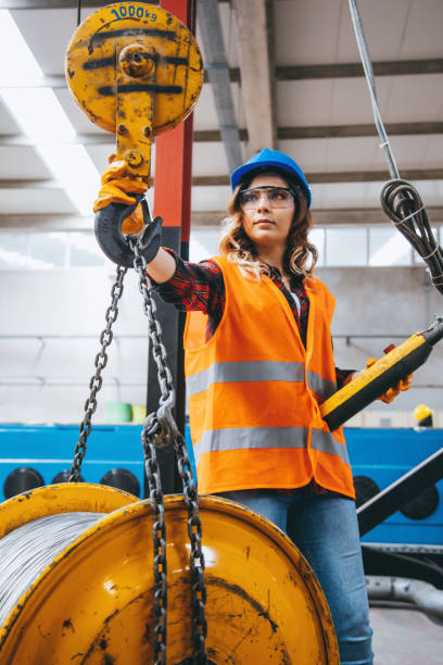 Young engineer woman holding and working with remote control for operating crane Skilled young heavy industry engineering worker woman holding crane hook button working with safety workwear and moving a massive metal construction object in warehouse of factory. XXXL size hoisting photos stock pictures, royalty-free photos & images