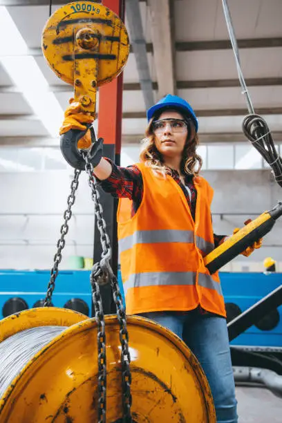 Skilled young heavy industry engineering worker woman holding crane hook button working with safety workwear and moving a massive metal construction object in warehouse of factory. XXXL size