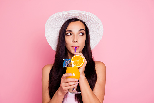 Close up studio photo portrait of thoughtful pondering with long brown hair lady holding orange beverage in hand wearing panama brimmed sunhat isolated bright pastel background