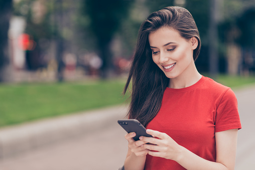 Girl in red tshirt with sweet smile writes on the phone a message about the place in the creek for a walk in the city on a summer day.