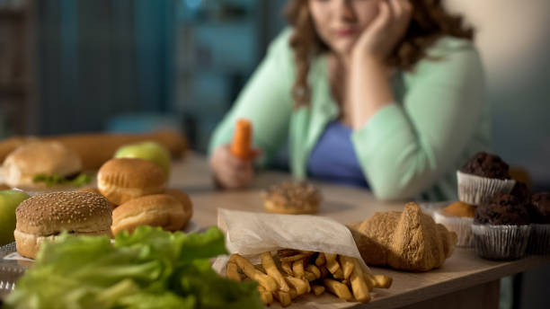 señora gorda deprimida sentada en la mesa llena de comida chatarra poco saludable, comer en exceso - comer demasiado fotografías e imágenes de stock