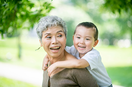 A grandmother and grandson are outdoors in a park. The grandson is climbing on his grandmother's shoulders.