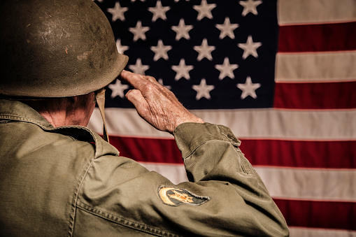 Senior adult man in military combat uniform saluting the American flag.  Rear view.