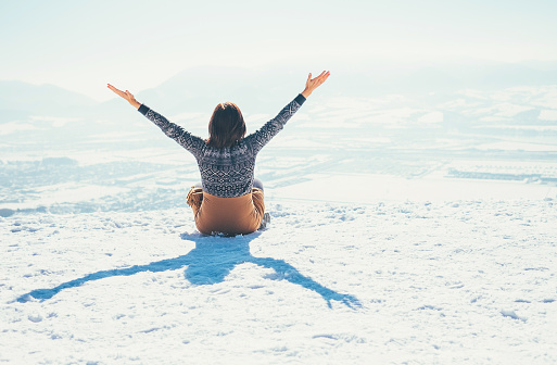 Happy woman sits on the snowy mountain hill over the city. Sunny winter day