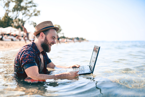 Bearded and handsome men with a brown straw hat  is cheating with his friends on water proof laptop in the water at the beach