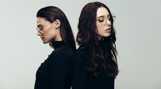 Two young woman in black outfit and glasses standing back to back. Two female models against grey background