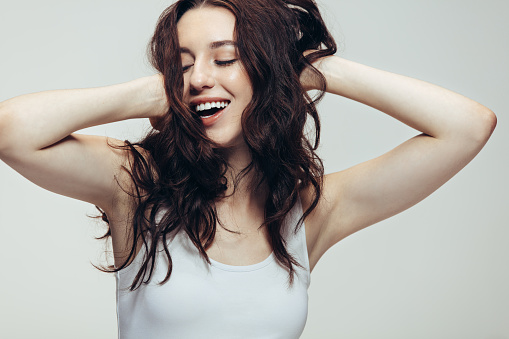 Woman in happy mood laughing with eyes closed and hands in hair. Close up of a young woman in casuals on grey background.