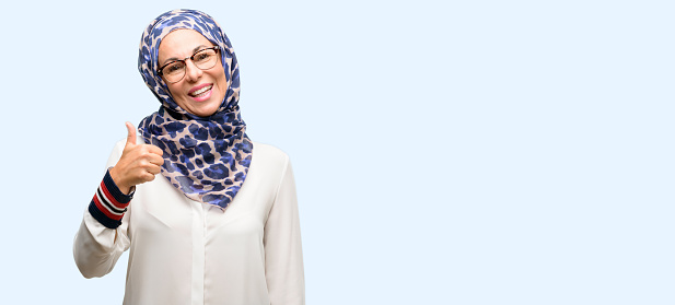 Middle age muslim arab woman wearing hijab smiling broadly showing thumbs up gesture to camera, expression of like and approval isolated blue background
