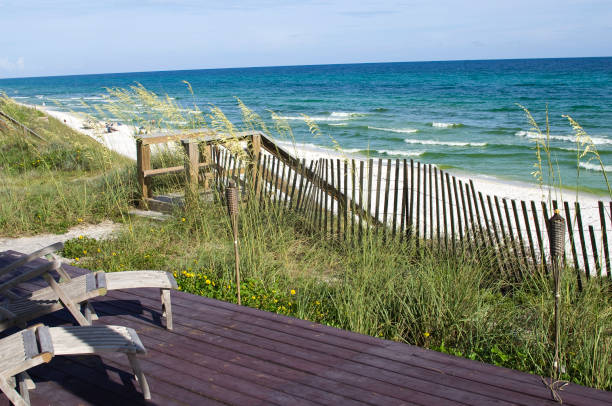 A peaceful beach with a white sand This color image if of beach front property with a beautiful view of the ocean. the picture includes a deck with deck chair next to the beach front with sand and ocean. this picture is a classic scenic beach front setting that is beautiful. there are also waves crashing against the beach. the mood is very peaceful and relaxing. the lighting is natural sunlight. and the background consists of the blue ocean and blue sky. pretty beach house stock pictures, royalty-free photos & images
