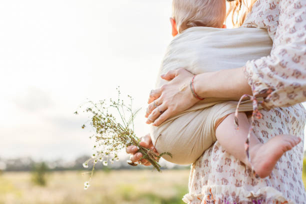 Little baby boy and his mother walking in the fields during summer day. Mother is holding and tickling her baby, babywearing in sling. Natural parenting concept stock photo