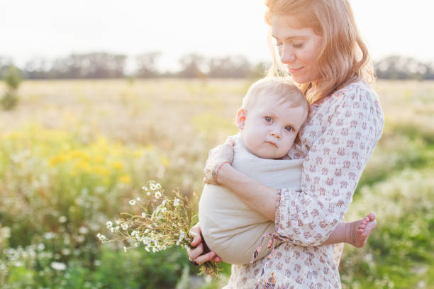 Little baby boy and his mother walking in the fields during summer day. Mother is holding and tickling her baby, babywearing in sling. Natural parenting concept stock photo