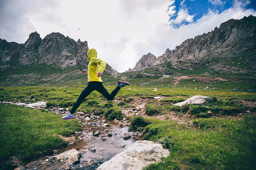 Woman trail runner jumping over samll river on beautiful mountains