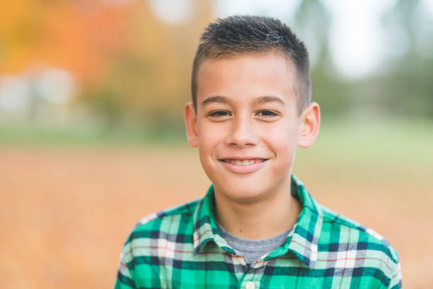 An elementary-age boy with braces smiles at the camera. The shot is a close-up of him as he stands in a city park in the fall leaves.
