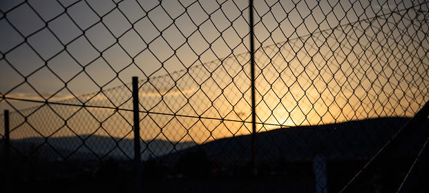 Steel wire mesh fence on a sunset background. Blurred city and mountains silhouette.
