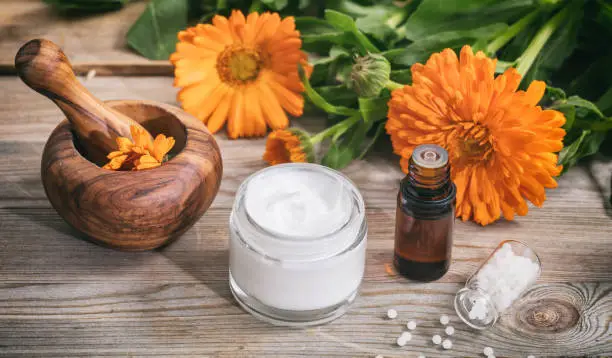 Calendula alternative medicine. Essential oil, ointment a mortar and white pills on a wooden table, fresh blooming calendula background,