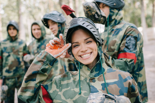 portrait of laughing female paintballer in uniform looking at camera while her team standing behind outdoors portrait of laughing female paintballer in uniform looking at camera while her team standing behind outdoors paintballing stock pictures, royalty-free photos & images