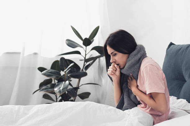 sick young woman sitting in bed and having cough while suffering from sore throat stock photo