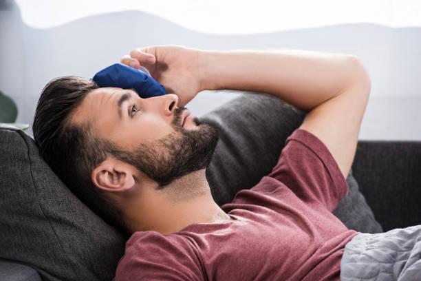 close-up portrait of sick young man lying on couch and holding ice pack on forehead - ice pack fotos imagens e fotografias de stock