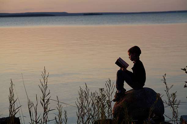 A boy reading a book outside on a porch overlooking the sea in warm late afternoon light.