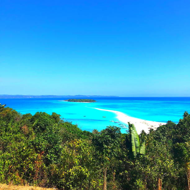 Walking on the sand towards the island you can see the water coming from both sides. \nIranja Island (Nosy Iranja) with palm trees and boats floating in the sea, Madagascar.\nSquare shape.