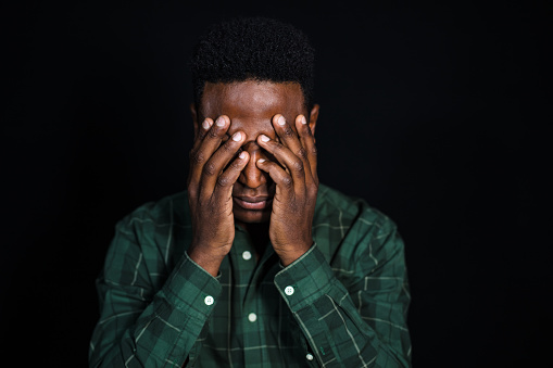 Portrait of african man with hand covering face and thinking. Male in checkered shirt looking worried on black background.