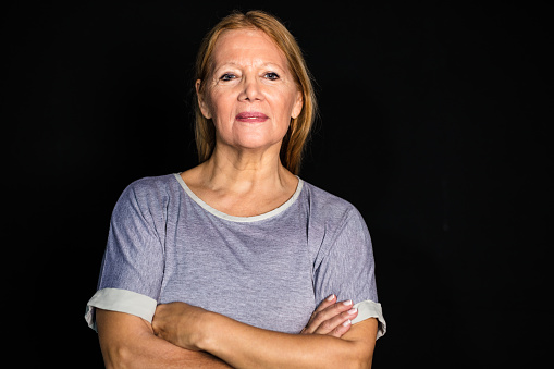 Portrait of senior woman against black background. Female wearing t-shirt standing with her arms crossed in studio.