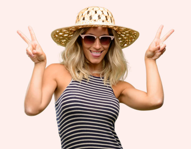 istock Young woman using sunglasses wearing summer hat looking at camera showing tong and making victory sign with fingers 1042437152
