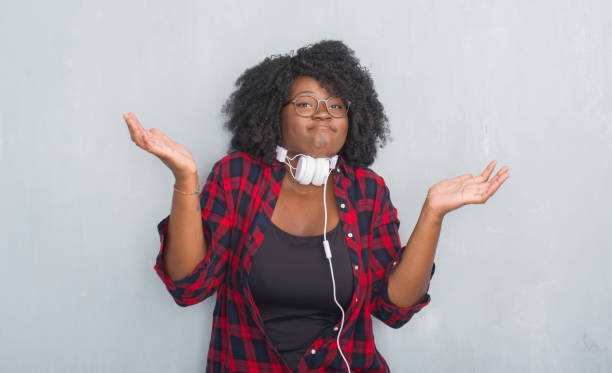 young african american woman over grey grunge wall wearing headphones clueless and confused expression with arms and hands raised. doubt concept. - head and shoulders audio imagens e fotografias de stock