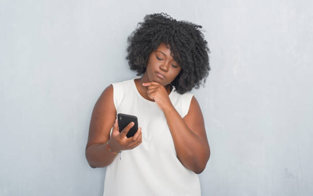 istock Young african american woman over grey grunge wall texting a message using smartphone serious face thinking about question, very confused idea 1042447528