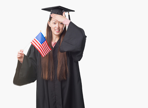 Young Chinese woman wearing graduate uniform holding United states of america flag stressed with hand on head, shocked with shame and surprise face, angry and frustrated. Fear and upset for mistake.