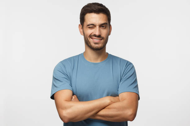 joven guapo en camiseta, con los brazos cruzados sonriendo y guiñando un ojo, mirando a cámara aislada sobre fondo gris - parpadear fotografías e imágenes de stock