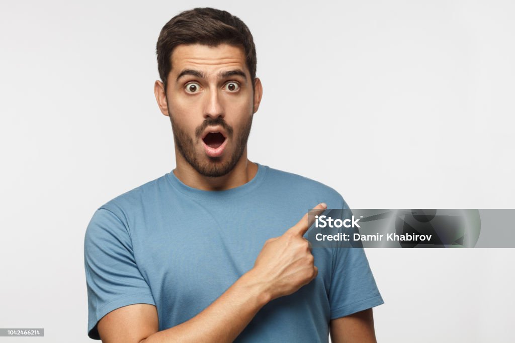 Young surprised man isolated on gray background in blue t-shirt looking at camera with open mouth, pointing right, copyspace for ads Surprise Stock Photo