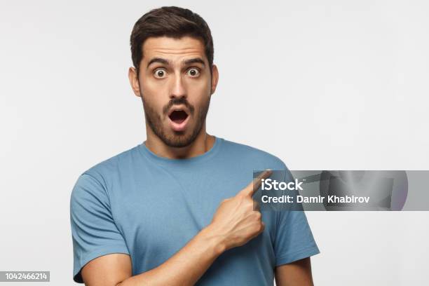 Young Surprised Man Isolated On Gray Background In Blue Tshirt Looking At Camera With Open Mouth Pointing Right Copyspace For Ads Stock Photo - Download Image Now