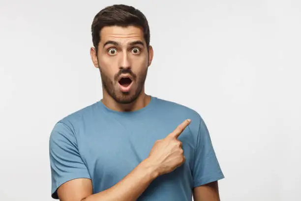 Photo of Young surprised man isolated on gray background in blue t-shirt looking at camera with open mouth, pointing right, copyspace for ads