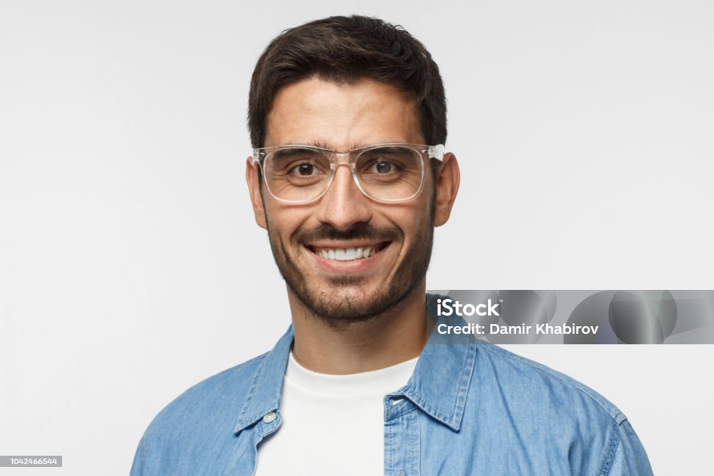 Jeune homme positif avec des lunettes, vêtu d’une chemise bleue clair et un t-shirt blanc, regardant la caméra avec un sourire heureux, isolé sur fond gris - Photo de Tête - Composition libre de droits