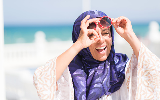 Middle age brunette arabian woman by the pool wearing sunglasses with happy face smiling doing ok sign with hand on eye looking through fingers