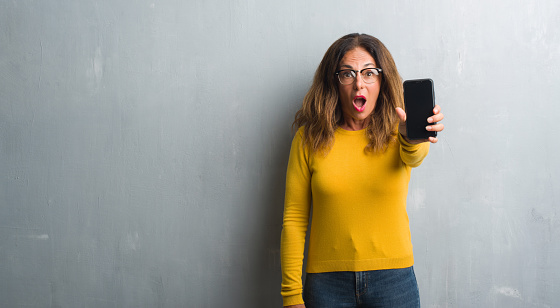 Middle age hispanic woman showing smarphone screen scared in shock with a surprise face, afraid and excited with fear expression