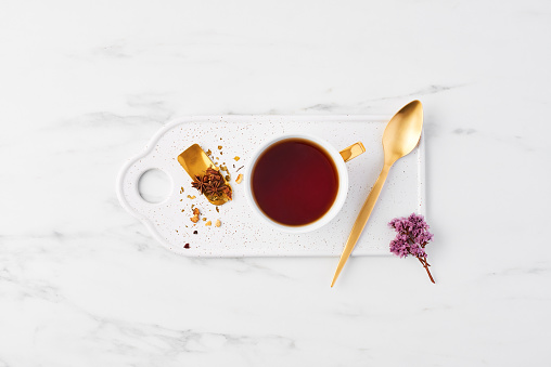 Top view of white cup of tea with herbs on tea mesure and golden spoon on serving plate. Cup of tea on white marble background with dried flowers. Flat lay.