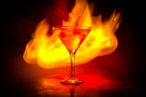 Martini in fire concept. Glass of famous cocktail Martini burning in fire at dark toned foggy background. Selective focus