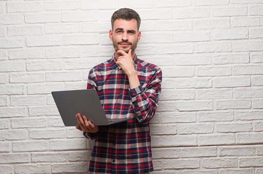 Young adult man over brick wall using laptop serious face thinking about question, very confused idea
